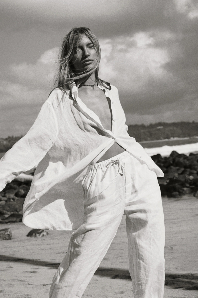 Black and white shot of a blonde woman in a linen shirt and pants posing on a beach with ocean and cloudy sky in the background.