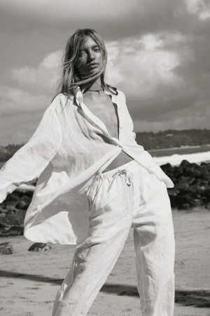 Black and white shot of a blonde woman in a linen shirt and pants posing on a beach with ocean and cloudy sky in the background.
