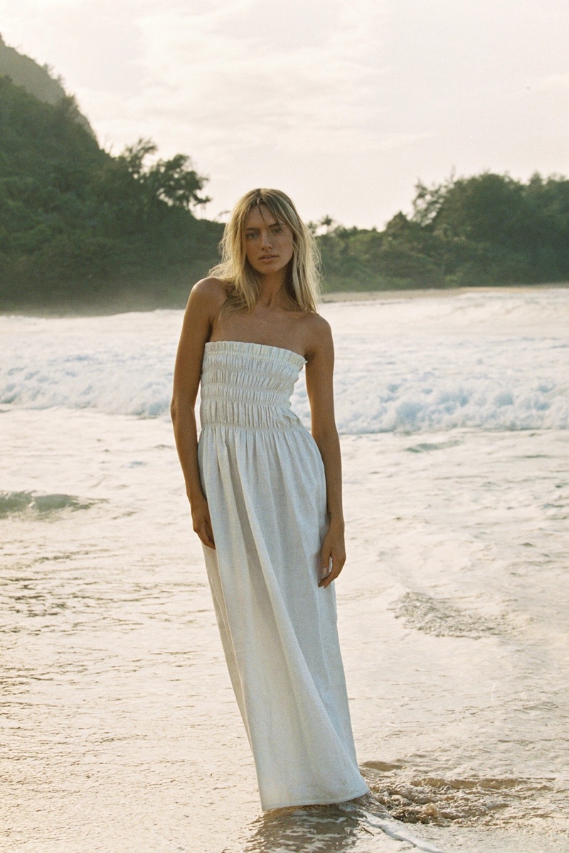 A woman stands on a sandy beach in a white strapless dress with ocean waves and a green, tree-covered hill in the background.