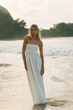 A woman stands on a sandy beach in a white strapless dress with ocean waves and a green, tree-covered hill in the background.