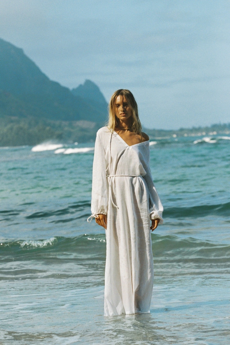 Model standing waist-deep in ocean water wearing a long white dress with mountains in the background