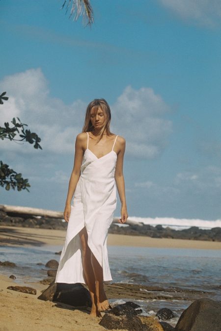 Model in white wrap dress walking along a sandy beach under a blue sky