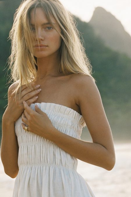 Blonde woman in white dress poses on beach with mountains in the background. Summer fashion editorial, beauty and nature portrait.