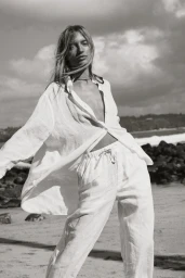 Model in a white linen shirt and matching pants posing on a sandy beach against a dramatic cloudy sky.