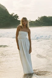 Maya Stepper in a white strapless maxi dress standing on a sandy beach with ocean waves for Sir The Label Holiday campaign.