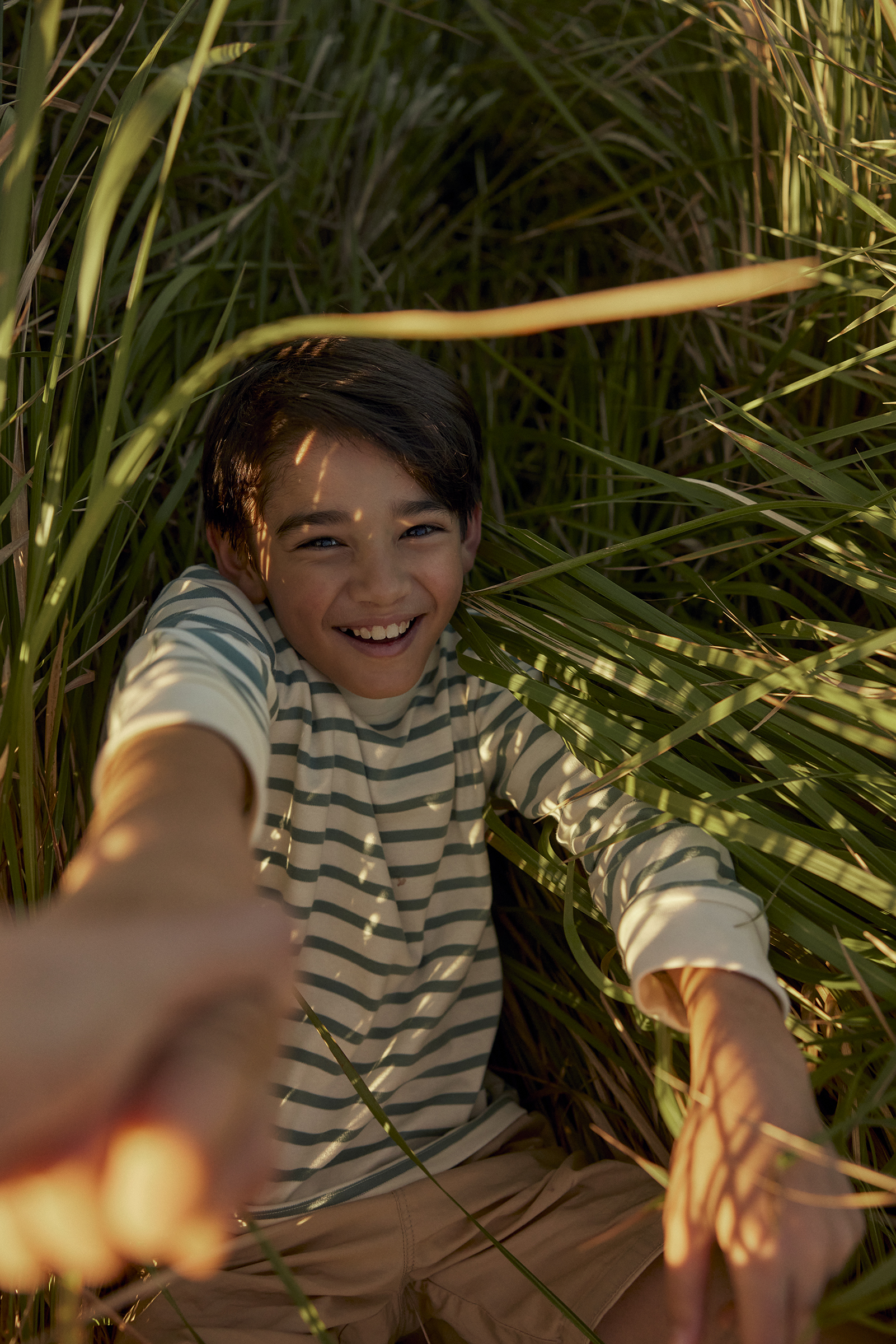 A smiling child with dark hair lies in tall green grass, looking up at the camera and holding a hand in a point-of-view outdoor shot.