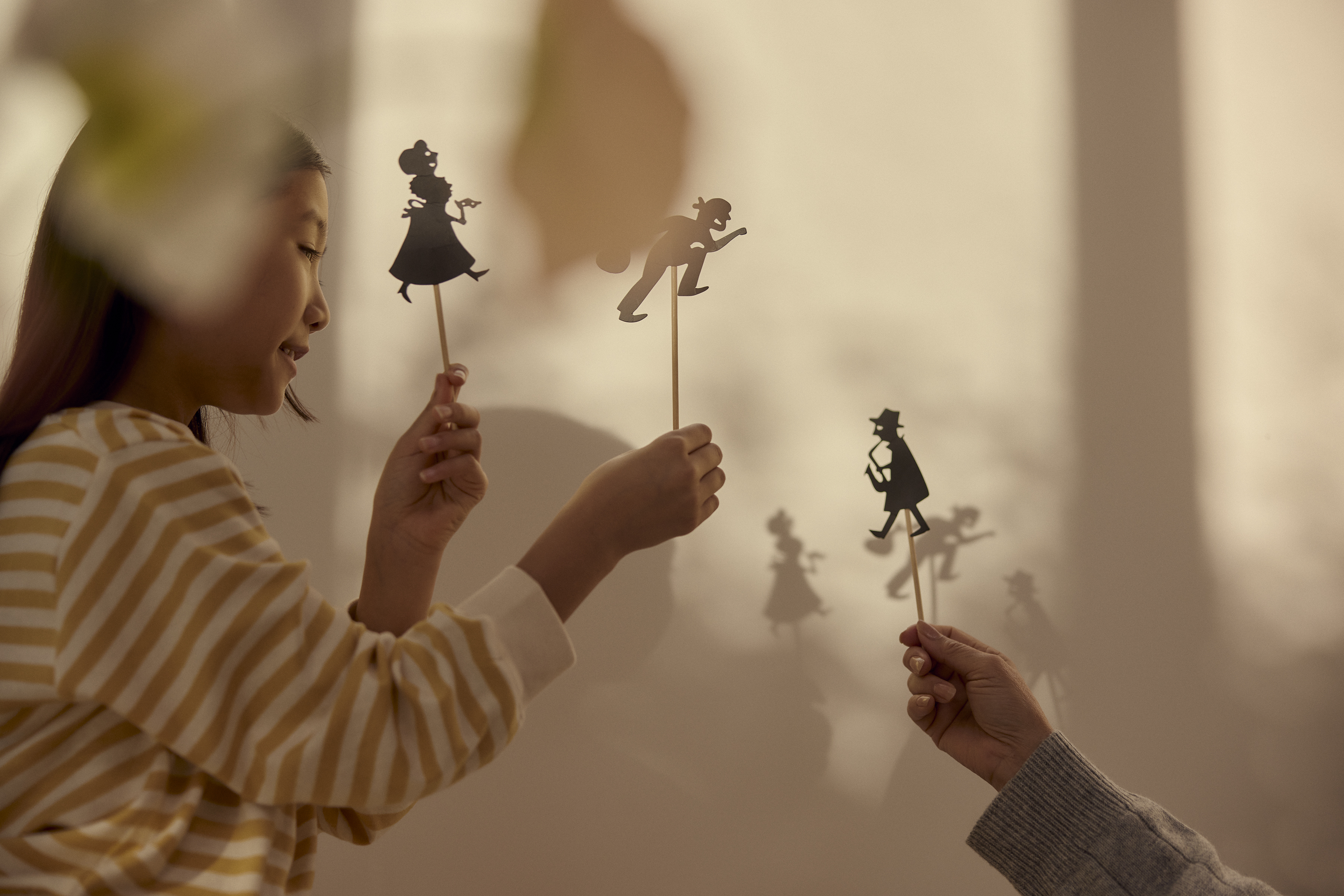 A child and an adult play with shadow puppets indoors against a beige wall, creating silhouettes of people in a vintage style.