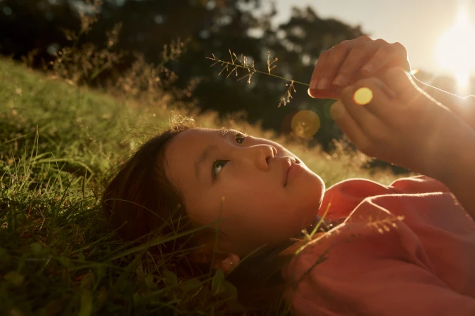 Young child lies in grassy field holding flower, looking up, lit by golden sun.