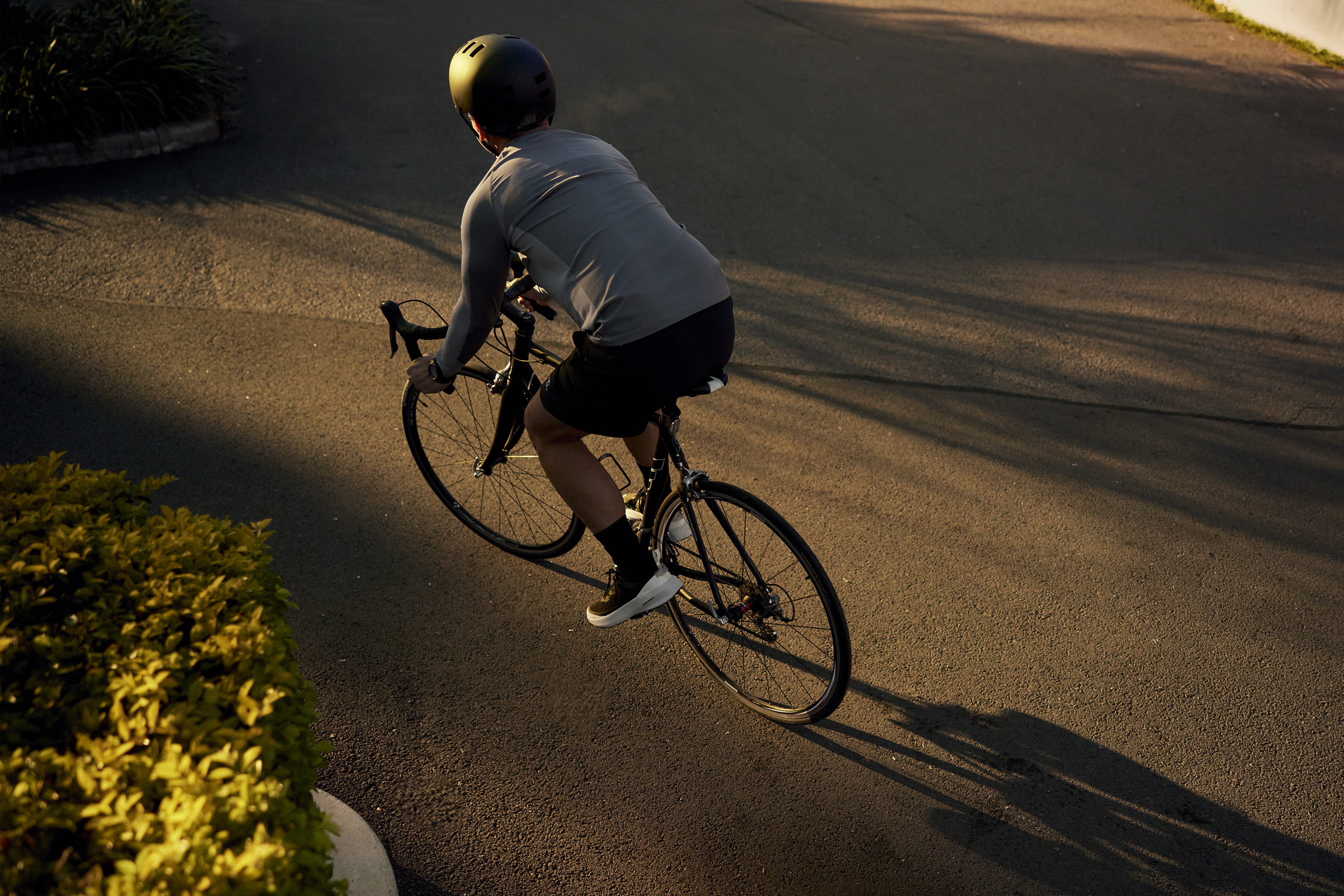 A cyclist rides a black road bike on an asphalt road under golden light. The rider wears a grey shirt and a dark helmet.