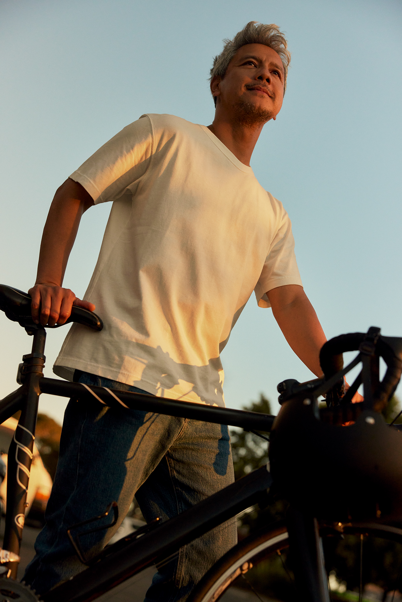 Man with grey hair wearing a white t-shirt and blue jeans standing outdoors next to a bicycle during sunset
