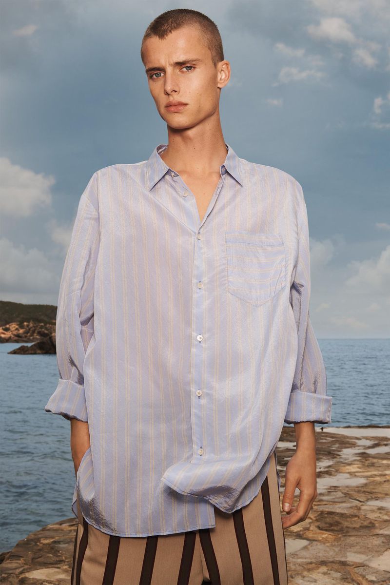 Portrait of a male model wearing a striped light blue collared shirt with brown striped trousers, posing outdoors near the ocean.