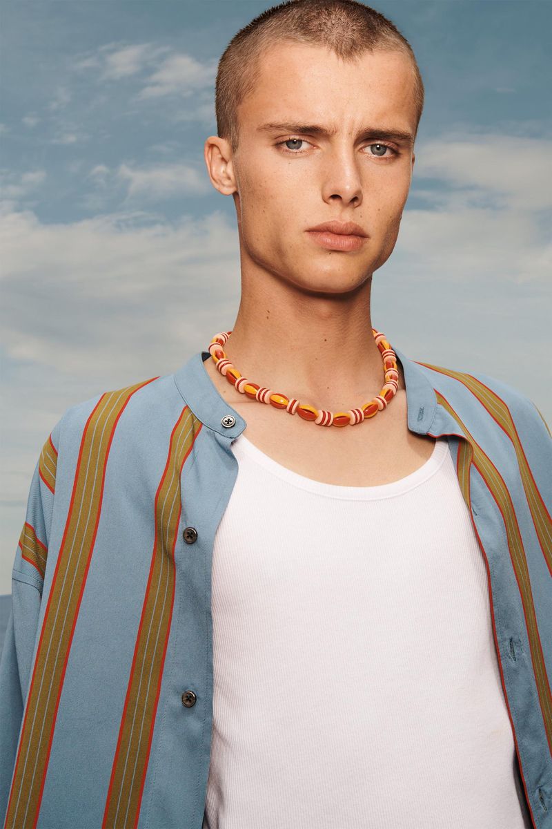 Portrait of a young adult man wearing a beaded necklace, white tank top, and blue striped button-down shirt against a cloudy sky.