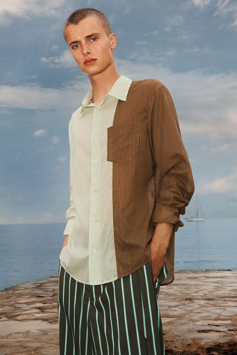 Adult male fashion model posing outdoors wearing a vertical-striped button-down shirt and striped pants against an ocean backdrop.