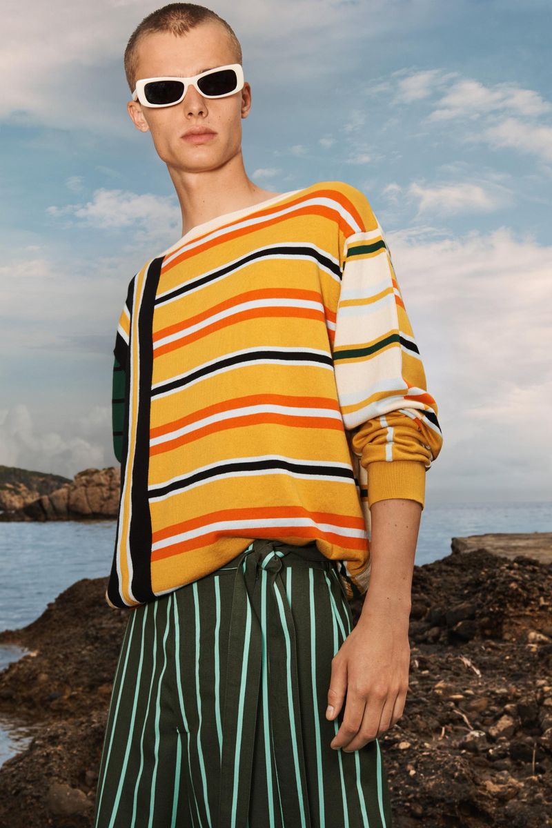 Man in striped sweater and striped pants poses outdoors with sea and rock formations in the background against a partly cloudy sky.