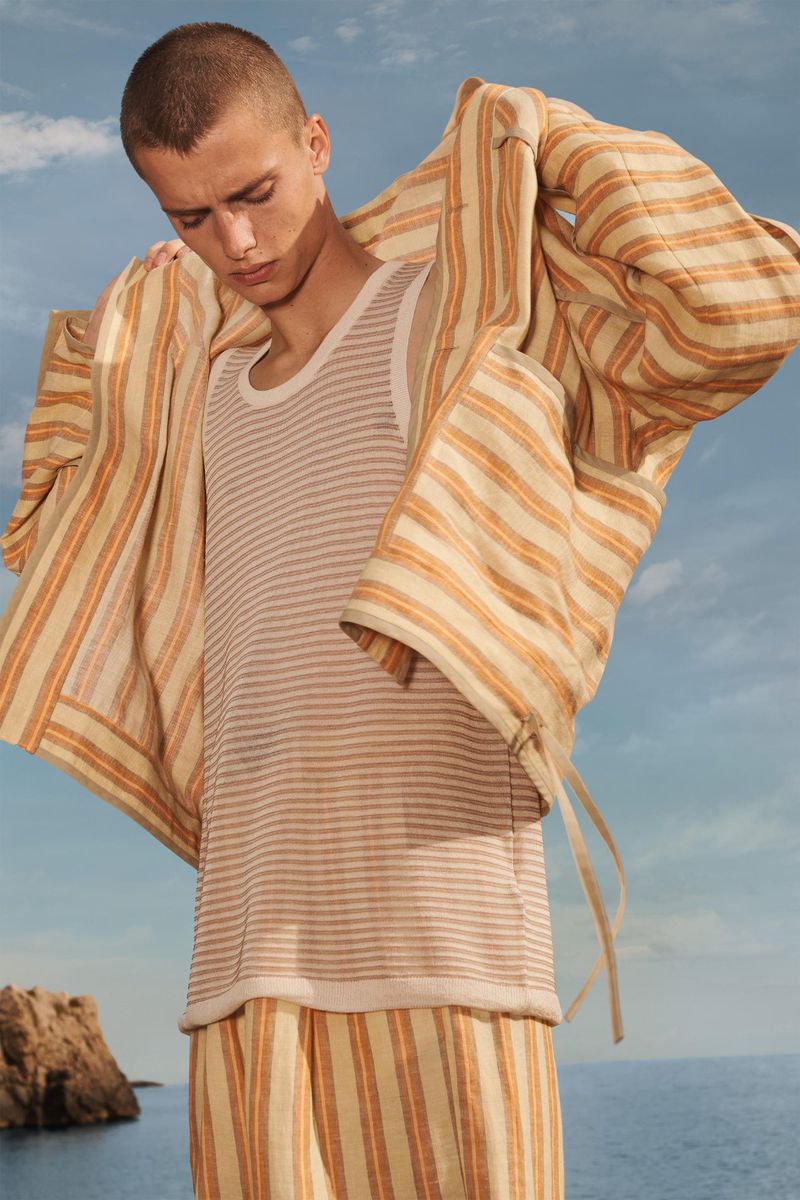 A young adult man in striped pajamas and robe poses outdoors with ocean and sky in the background. Fashion shot, neutral colors.