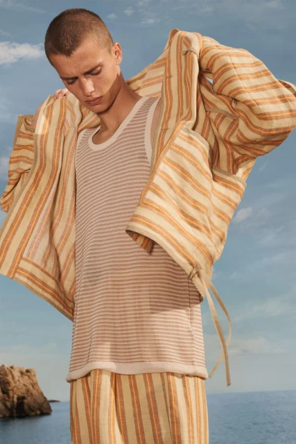 A young adult man in striped pajamas and robe poses outdoors with ocean and sky in the background. Fashion shot, neutral colors.