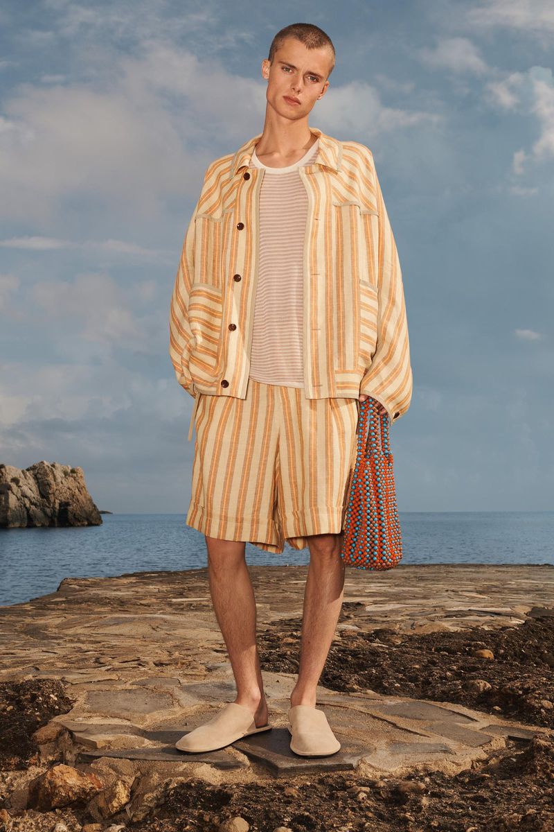 Male fashion model posing on a stone surface wearing a striped linen jacket, matching shorts, a white tee shirt, and slip-on shoes.