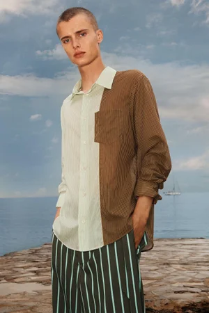 Male model wearing a two-tone striped shirt and striped trousers standing on a stone pier by the sea under an open sky.