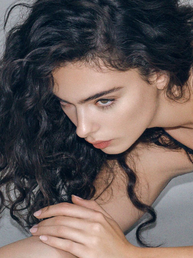 Close-up portrait of a young adult female model with dark curly hair, fair skin, and a serene expression, posing indoors in a photo studio.
