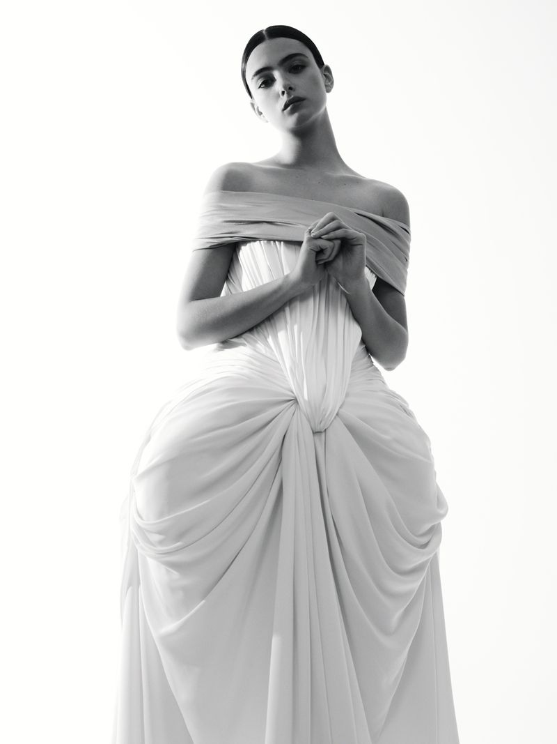 Monochrome fashion portrait of a young woman in an off-the-shoulder gown with draped fabric, hands clasped, against a white backdrop.