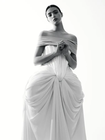 Monochrome fashion portrait of a young woman in an off-the-shoulder gown with draped fabric, hands clasped, against a white backdrop.