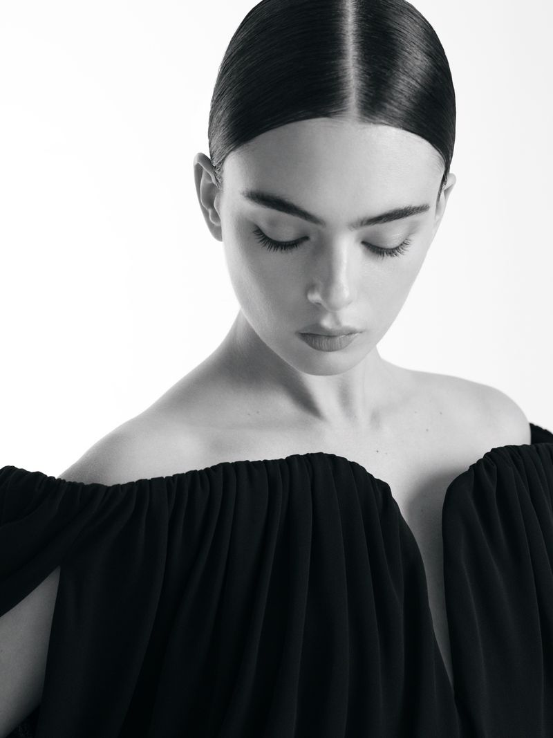 Black and white portrait of a woman with dark hair pulled back, wearing a dark dress with an off-the-shoulder neckline. Studio lighting.