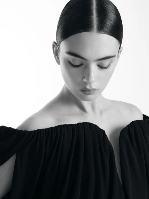 Black and white portrait of a woman with dark hair pulled back, wearing a dark dress with an off-the-shoulder neckline. Studio lighting.