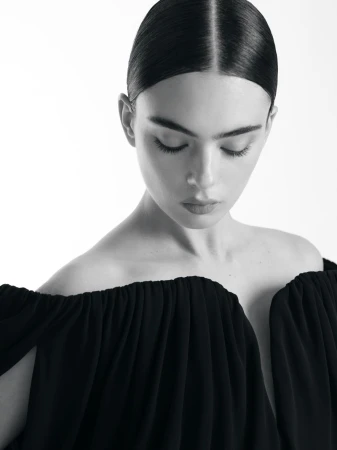 Black and white portrait of a woman with dark hair pulled back, wearing a dark dress with an off-the-shoulder neckline. Studio lighting.
