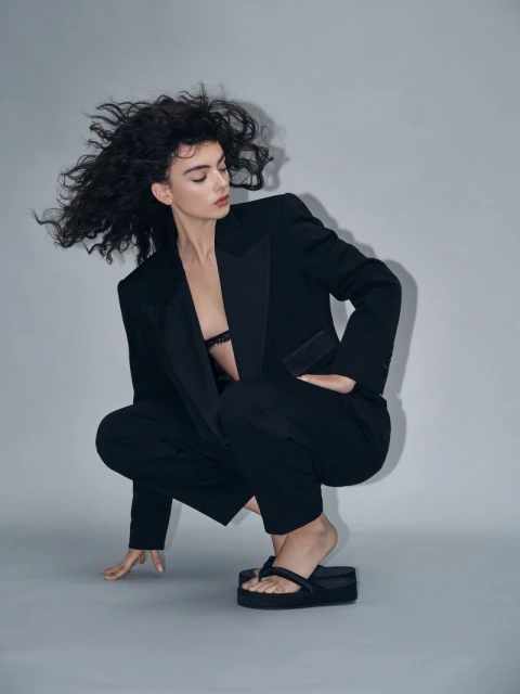 A woman with curly dark hair poses in a black blazer and pants with black flip-flops, squating in a photo studio with a neutral background.