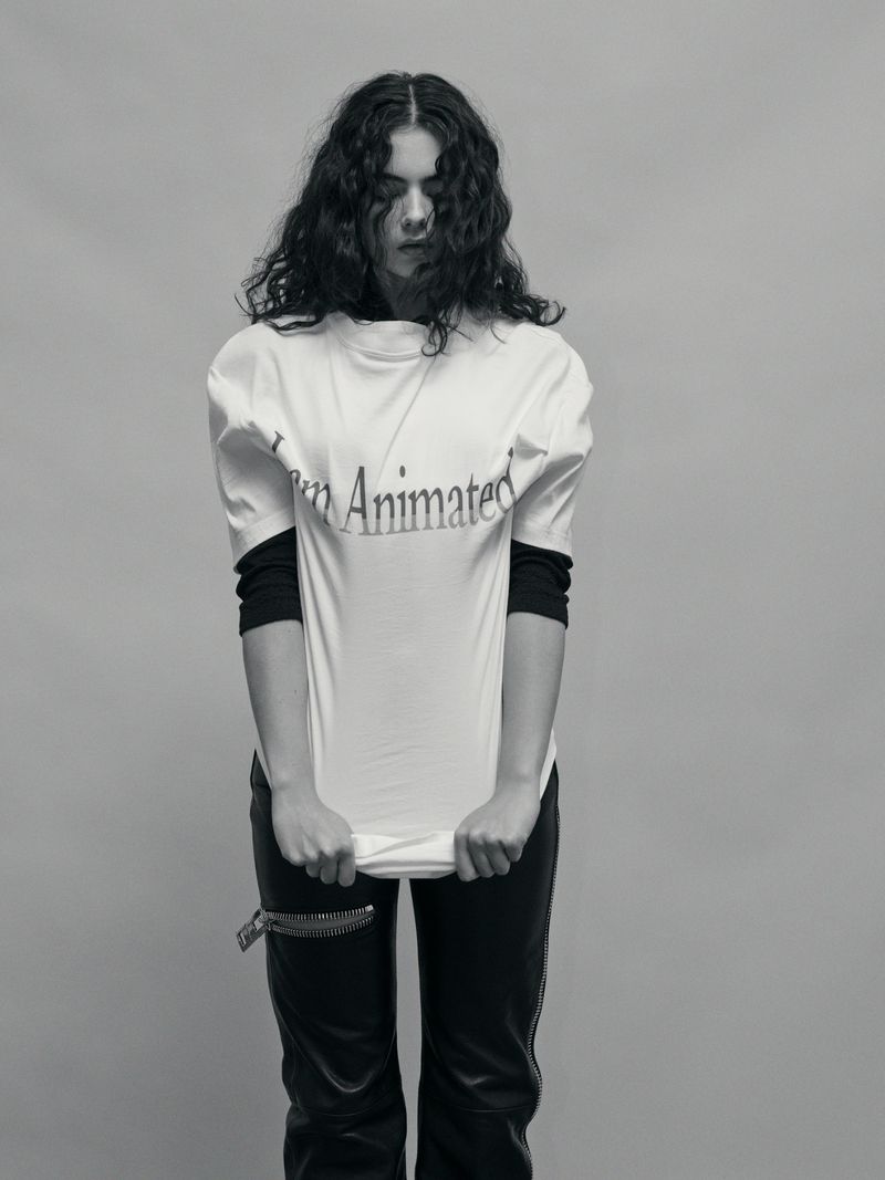 Monochrome portrait of a young woman with curly hair wearing a 'I am Animated' t-shirt and holding up the bottom of the shirt.