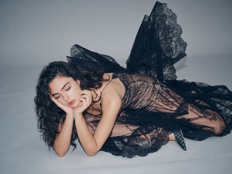 Woman in a black lace dress lying on a grey floor with her head propped up by her hands in a studio setting. Fashion shot.