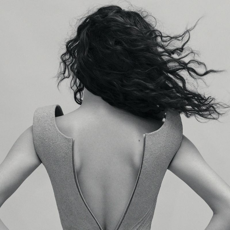 Back view of adult female with dark wavy hair in a light grey structured dress with a metal zipper, posing in a neutral grey studio.