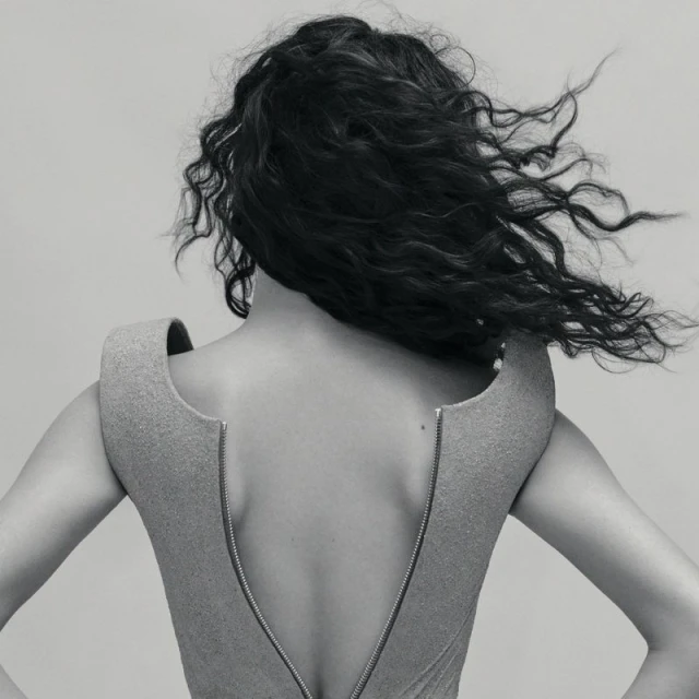 Back view of adult female with dark wavy hair in a light grey structured dress with a metal zipper, posing in a neutral grey studio.