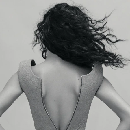 Back view of adult female with dark wavy hair in a light grey structured dress with a metal zipper, posing in a neutral grey studio.