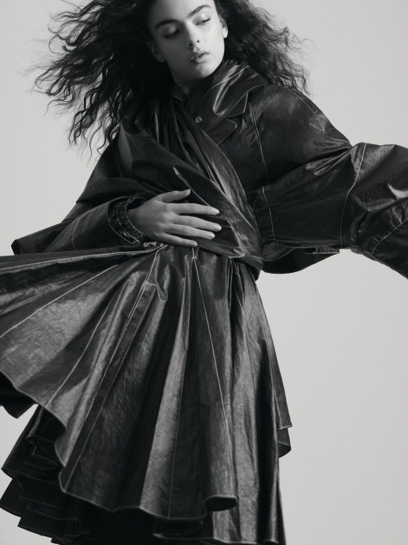 Monochrome portrait of a young woman in a dark, flowing dress, with expressive, voluminous hair, and soft, diffused lighting in studio.