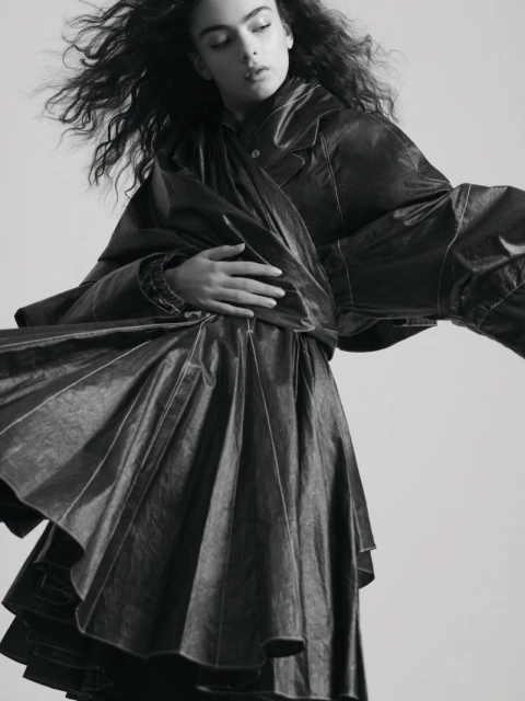 Monochrome portrait of a young woman in a dark, flowing dress, with expressive, voluminous hair, and soft, diffused lighting in studio.