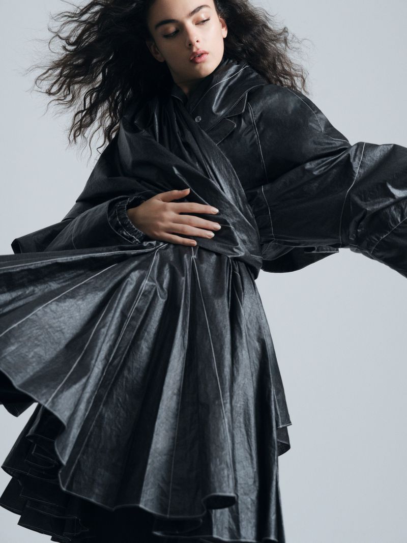 Deva Cassel posing in a voluminous black draped leather coat with windswept hair against a light grey studio background.