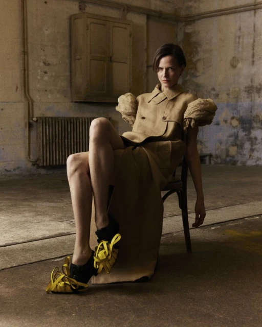 A woman seated on a chair, dressed in a beige, oversized suit-dress and yellow ribbon shoes, poses in a distressed, industrial-style indoor setting.
