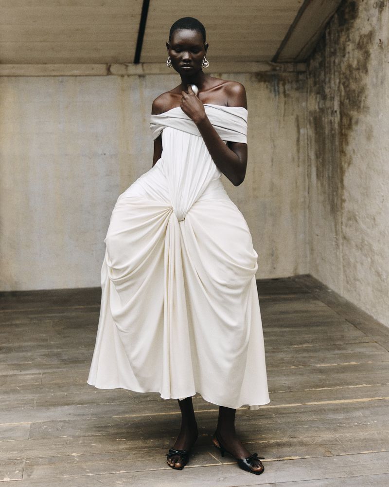 Woman in an off-the-shoulder cream-colored gown posing in front of a textured wall and floor, accessorized with earrings and rings.