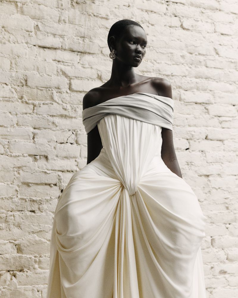 A model with short dark hair wearing a draped white off-the-shoulder gown standing against a textured white brick wall.
