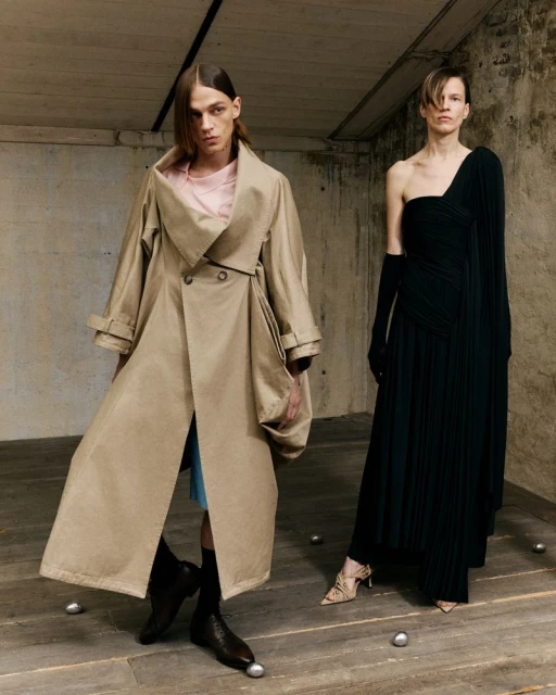 Two fashion models in a neutral setting. One wears a draped black dress and the other a trench coat, posing expressively.