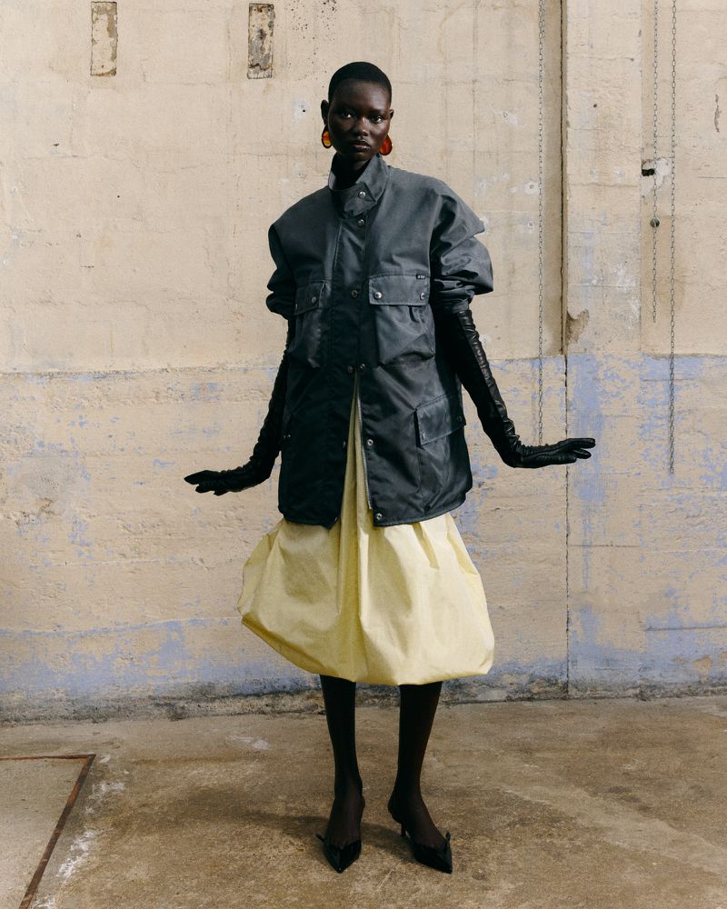 Fashion shot of a Black female model in a grey jacket and yellow skirt, standing in a photo studio with painted concrete walls.