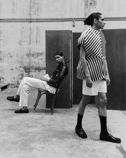 Two fashion models pose indoors: one sits on a chair in a leather jacket and white pants, while the other stands in a striped top.