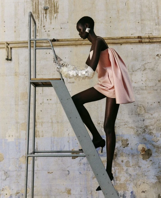 Fashion editorial of an African-American adult female model wearing a light pink mini dress and sheer gloves, climbing a metal ladder.