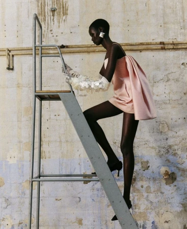 Fashion editorial of an African-American adult female model wearing a light pink mini dress and sheer gloves, climbing a metal ladder.