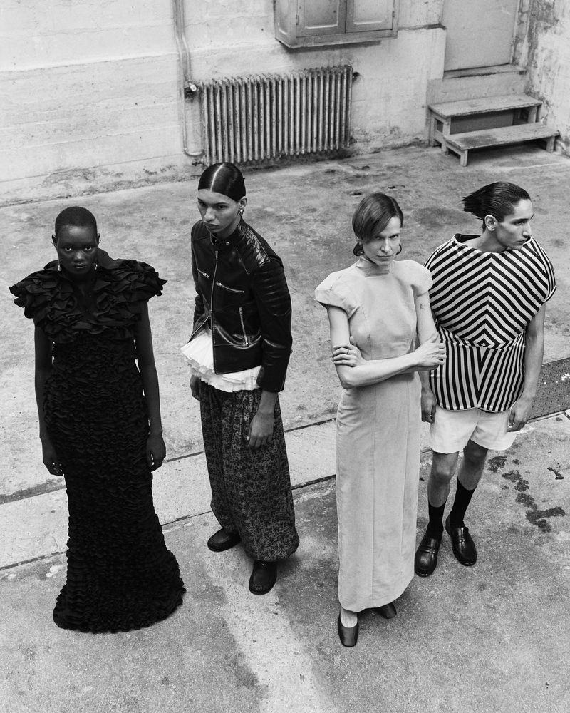 Four fashion models in edgy designs pose outdoors; a black ruffled dress, a leather jacket and patterned skirt, a long dress, and striped top.