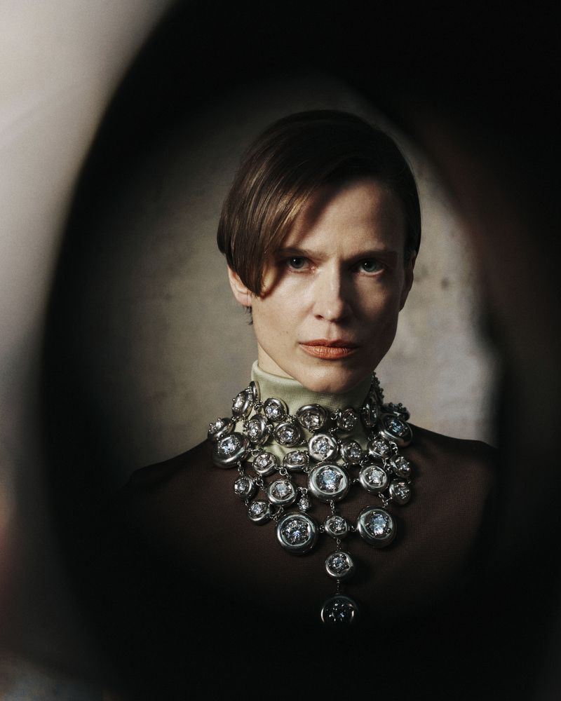 Portrait of a brunette adult woman wearing a large ornate silver necklace with clear gemstones, shot through a dark oval frame.