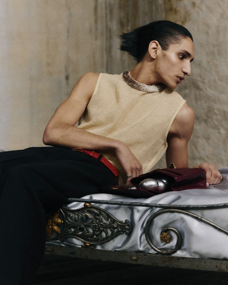 Fashion shot of a person wearing a beige tank top and black pants sitting on a silver bed in an indoor location with neutral tones.