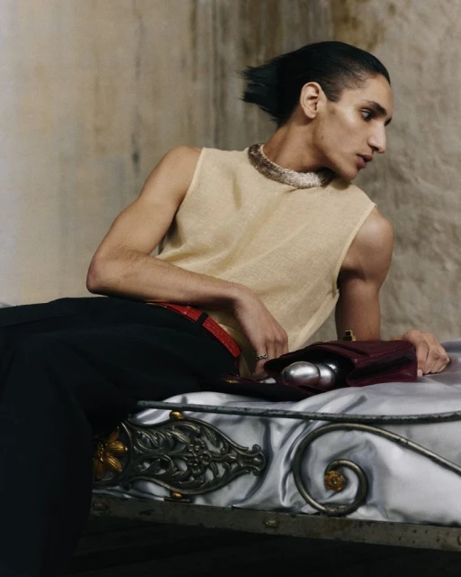 Fashion shot of a person wearing a beige tank top and black pants sitting on a silver bed in an indoor location with neutral tones.