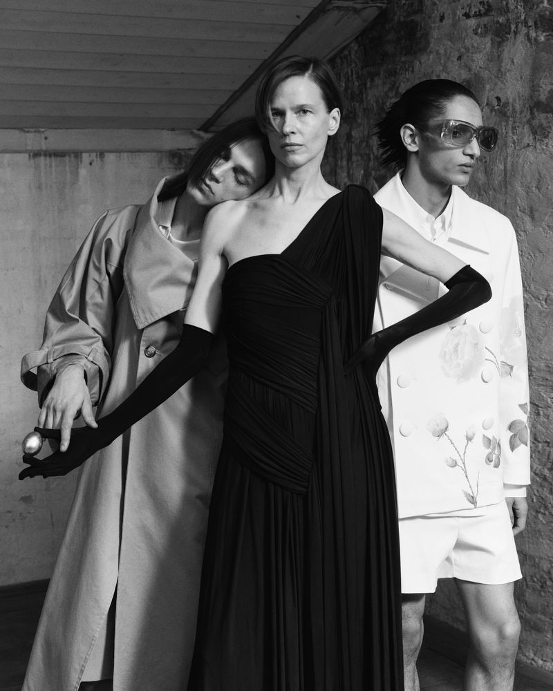 Three people in a black and white fashion shot. A woman in a black gown is centered between two others in a studio environment.